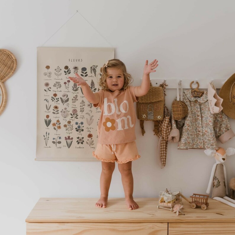 Op de afbeelding: Een jong kind draagt een perzikkleurige t-shirt en short set met het woord "bloom" en een bloemenafbeelding. Een botanische print hangt aan de muur achter het kind. Een kleine rugzak en jas zijn ook zichtbaar.