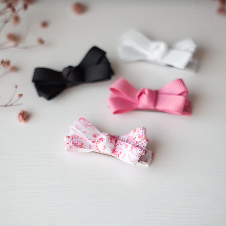 May include: Four hair bows in different colors: black, white, pink, and a floral print. The bows are made of a grosgrain ribbon and are arranged on a white surface. The bows are small and delicate, suitable for various hairstyles.