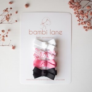 May include: A set of five hair bows in various colors, including white, pink, and black, arranged on a white card. The bows are made of ribbon and feature a classic bow shape. The card has the text "bambi lane hair accessories" printed on it.