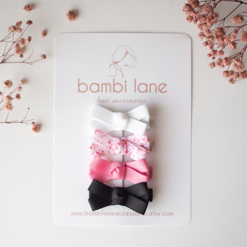May include: A set of five hair bows in various colors, including white, pink, and black, arranged on a white card. The bows are made of ribbon and feature a classic bow shape. The card has the text "bambi lane hair accessories" printed on it.