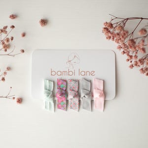 May include: A set of five assorted hair clips in pastel colors, including mint green, pink, and gray, displayed on a white card with the "bambi lane" logo. The clips feature a bow design and are suitable for various hairstyles.