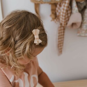 Op de afbeelding: Een licht perzikkleurige strik haarclip is bevestigd aan de zijkant van het blonde haar van een persoon. De strik is gemaakt van een zacht, getextureerd materiaal. De achtergrond is een lichtgekleurde muur met een houten plank en hangende items.