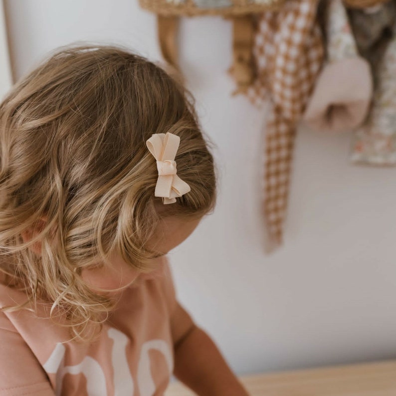 Op de afbeelding: Een licht perzikkleurige strik haarclip is bevestigd aan de zijkant van het blonde haar van een persoon. De strik is gemaakt van een zacht, getextureerd materiaal. De achtergrond is een lichtgekleurde muur met een houten plank en hangende items.