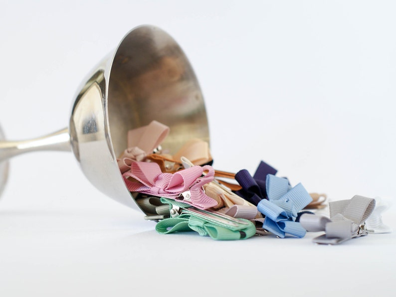 Op de afbeelding: Een zilveren beker die is omgevallen en een verzameling kleurrijke lint haarclips uitstort. De clips zijn in verschillende pastelkleuren, waaronder roze, blauw en beige. De achtergrond is effen wit.