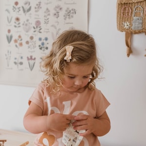 Op de afbeelding: Een jong kind draagt een perzikkleurig T-shirt met witte letters en een perzikkleurige haarclip met strik. Het kind houdt een kleine doek met patroon vast. Op de achtergrond is een bloemen botanische print te zien.