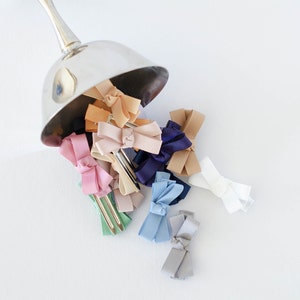 Op de afbeelding: Een verzameling kleurrijke haarstrikken die uit een zilveren bakje vallen. De strikken zijn in verschillende tinten, waaronder roze, blauw, beige en wit. Elke strik is bevestigd aan een metalen haarclip, wat een charmant en stijlvol accessoire cre&euml;ert.