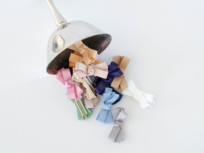 Op de afbeelding: Een verzameling kleurrijke haarstrikken die uit een zilveren bakje vallen. De strikken zijn in verschillende tinten, waaronder roze, blauw, beige en wit. Elke strik is bevestigd aan een metalen haarclip, wat een charmant en stijlvol accessoire cre&euml;ert.