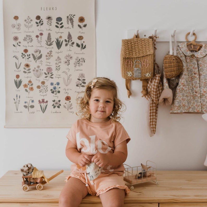 Op de afbeelding: Een kind draagt een perzikkleurig T-shirt en korte broek met de tekst "blo" erop. Een poster met bloemen hangt aan de muur. Een kleine houten wagen en een metalen speelgoedwagen staan op het houten oppervlak. Een rieten rugzak hangt aan de muur.