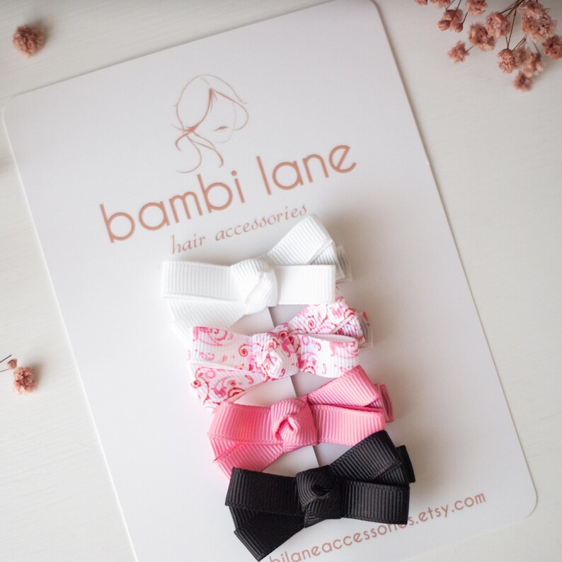 May include: Four small hair bows in white, pink, and black, arranged on a white card. One bow is white, one is pink with a floral pattern, one is solid pink, and one is black. The card has the text "bambi lane hair accessories" and a website address.