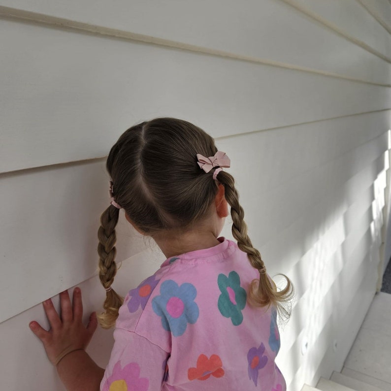 Op de afbeelding: Een kind met blond haar in twee vlechten, versierd met roze strikken. Het kind draagt een roze t-shirt met een bloemenpatroon in blauw, groen en oranje. Het kind kijkt naar een witte muur.