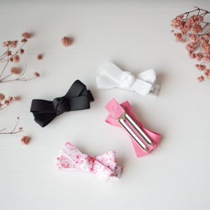 May include: Four small hair bows in black, white, pink, and a patterned pink and white design. The bows are made of ribbon and attached to metal clips. The bows are arranged on a white surface with dried flowers.