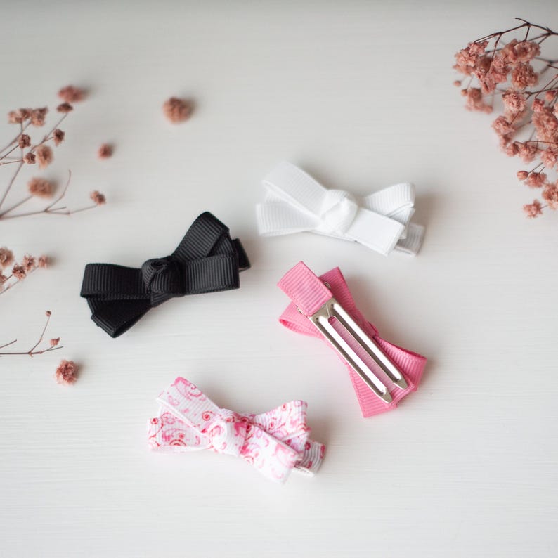 May include: Four small hair bows in black, white, pink, and a patterned pink and white design. The bows are made of ribbon and attached to metal clips. The bows are arranged on a white surface with dried flowers.