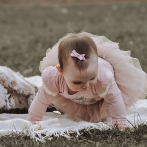 May include: A baby wearing a pink long-sleeved bodysuit and a matching tutu crawling on a white blanket. A small pink bow adorns the baby's hair. The scene is set outdoors on a grassy surface, with a faux fur pillow in the background.