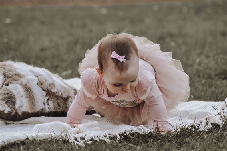 May include: A baby wearing a pink long-sleeved bodysuit and a matching tutu crawling on a white blanket. A small pink bow adorns the baby's hair. The scene is set outdoors on a grassy surface, with a faux fur pillow in the background.