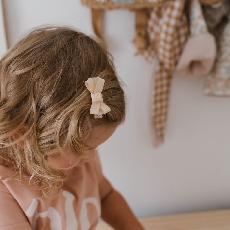 May include: A small, light peach-colored bow hair clip is attached to the side of a child's blonde hair. The bow is made of a soft fabric and is secured with a metal clip. The background is a blurred view of a wall and some hanging items.