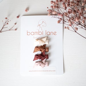 May include: Set of five mini hair bows in shades of brown, beige, and pink. The bows are arranged vertically on a white card with the text "bambi lane hair accessories" and a website address.