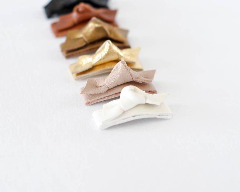 May include: A set of eight leather hair clips in various colors, including black, brown, gold, and white. The clips are arranged in a row, with the white clip at the bottom.