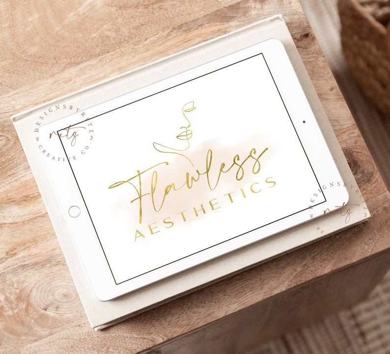 May include: A white tablet screen displaying a gold logo with the text "Flawless Aesthetics" and a line drawing of a face. The logo is on a light brown watercolor background.