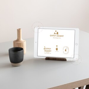 May include: A white tablet displaying a logo for a business called "Gypsy Desert Boutique Co." The logo features a stylized cactus and the text "Gypsy Desert Boutique Co." in a gold color. The tablet is on a wooden stand and a wooden vase with a black mug is on a white table.