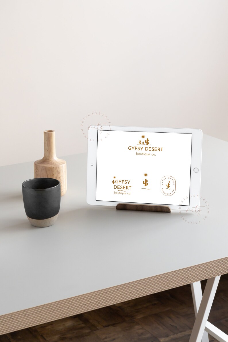May include: A white tablet displaying a logo for a business called "Gypsy Desert Boutique Co." The logo features a stylized cactus and the text "Gypsy Desert Boutique Co." in a gold color. The tablet is on a wooden stand and a wooden vase with a black mug is on a white table.