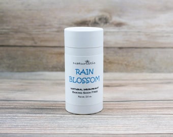 May include: White deodorant stick with the text "Naturistc Rain Blossom Natural Deodorant Baking Soda Free Net wt. 2.6 oz."