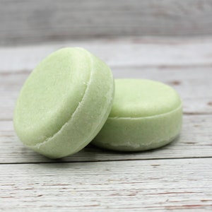 May include: Two round, green solid shampoo bars on a white wooden surface.