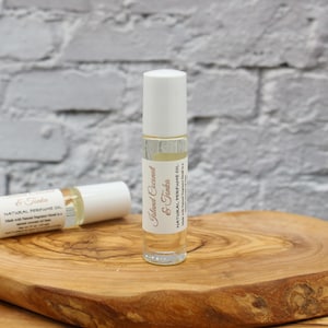 May include: A clear glass roller bottle with a white cap and a white label that reads "Island Coconut & Tonka" and "Natural Perfume Oil". The bottle is sitting on a wooden surface.