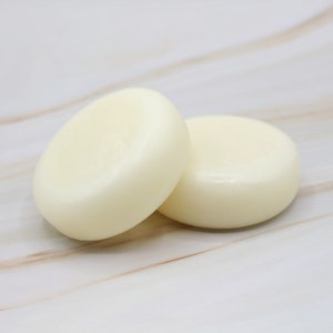 May include: Two round, white, solid soap bars on a white marble surface.