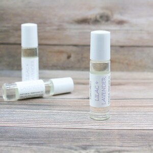 May include: Three glass roller bottles with white caps and labels. The label on the front bottle reads "Lilac & Lavender Natural Perfume Oil".