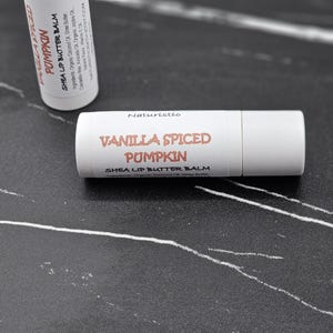 May include: Two white cylindrical lip balm tubes with labels reading Vanilla Spiced Pumpkin Shea Lip Butter Balm and Naturistic. The labels list ingredients including organic coconut oil, shea butter, candelilla wax, avocado oil, and jojoba oil.