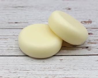 May include: Two round, pale yellow soap bars on a white wooden surface.