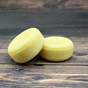May include: Two round, pale yellow solid shampoo bars. One bar is leaning against the other, which is upright. The bars have a smooth, slightly domed top and a flat bottom. The background is a dark wood surface.