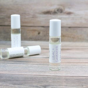 May include: Three white roller bottles with clear liquid inside. The bottle in the center has a label that reads "Lilac + Lavender Natural Perfume Oil".
