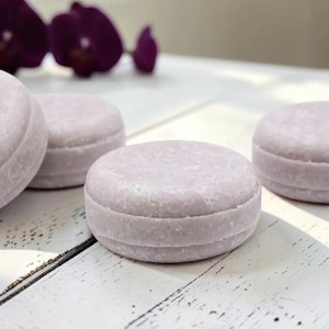 May include: Three round, light lavender solid shampoo bars with a smooth, matte texture and a distinct horizontal groove around the center. The bars are arranged in a cluster, showcasing their uniform circular shape and pale purple color.