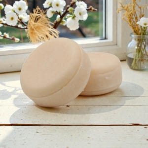 May include: Two round, light beige soap bars are displayed on a white wooden surface. The soaps are smooth and have a slightly rounded edge. The background includes a window with white flowers and a small glass vase with flowers.