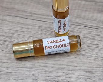 May include: Two clear glass rollerball perfume bottles with gold caps containing amber-colored liquid. The white labels feature the text Vanilla Patchouli and Natural Perfume Oil in brown lettering, with one bottle standing upright and one lying horizontally.