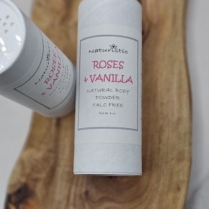 May include: Two white cylindrical containers of natural body powder, one upright and one tilted. The label reads "Roses & Vanilla" and "Talc Free." The net weight is 4 oz. The containers are on a wooden surface.