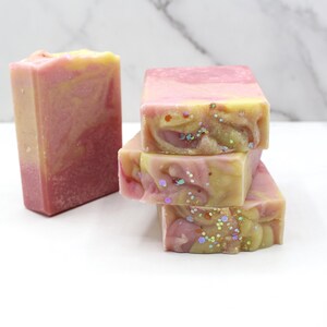 May include: A stack of three pink and yellow swirled soaps with iridescent glitter, next to a single bar of soap. The soaps have a marbled appearance and are on a white surface.