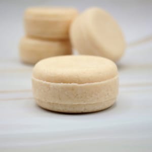 May include: Close-up of a single round, off-white solid shampoo bar on a white surface. Three more shampoo bars are out of focus in the background.