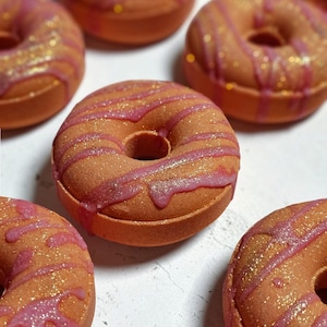 May include: A collection of round, donut-shaped bath bombs in a warm orange hue. Each bath bomb features a textured surface with a pink, drizzled icing pattern and a dusting of gold glitter particles across the top and sides of the circular form.