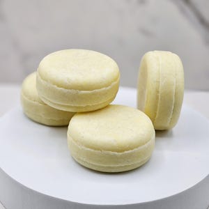 May include: Four pale yellow, round shampoo bars are stacked on a white, circular platform. The bars have a layered appearance, with visible ridges around the sides. The overall aesthetic is clean and minimalist, suggesting a natural or eco-friendly product.