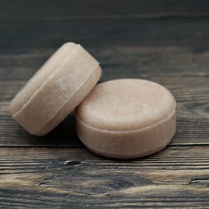 May include: Two round, light pink shampoo bars on a wooden surface.