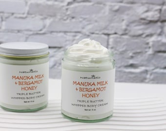 Può includere: Due barattoli di vetro di crema corpo montata con coperchi bianchi. Entrambi i barattoli hanno un'etichetta bianca con il testo "Naturistio Manuka Milk + Bergamot Honey Triple Butter Whipped Body Cream Net wt. 6 oz."