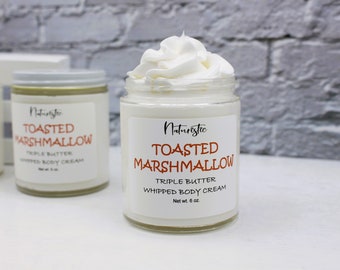 May include: Two glass jars of body cream with white labels and silver lids. The labels read "Naturistic TOASTED MARSHMALLOW TRIPLE BUTTER WHIPPED BODY CREAM Net wt. 6 oz." One jar is topped with a swirl of white cream.