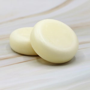 May include: Two round, white, solid soap bars on a light brown marble surface.