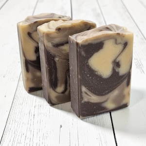 May include: Three rectangular bars of handmade soap with a marbled design. The soap features swirls of brown, tan, and cream colors. The bars are arranged on a white wooden surface, showcasing the natural tones and textures of the soap.