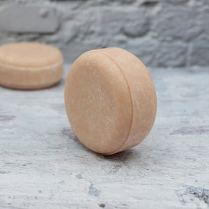 May include: A round, light brown solid shampoo bar on a white textured surface.