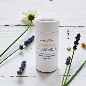 May include: A white deodorant stick with the text "Chamomile + Lavender" and "Baking Soda Free." The product is surrounded by lavender sprigs, a daisy, and yellow flower petals. Net weight is 2.6 oz.