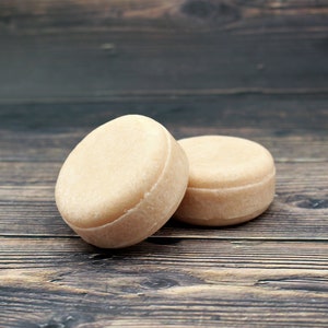 May include: Two round, light brown shampoo bars on a wooden surface.