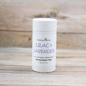 May include: A white deodorant stick with the text "Lilac & Lavender" in purple script. The label also reads "All Natural Deodorant Baking Soda Free" and "Net wt. 2.6 oz.". The product is on a light wood-grain surface.